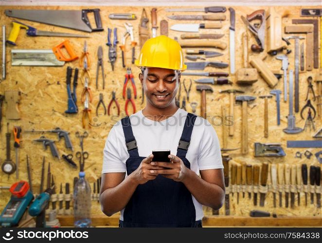 profession, construction and building - happy smiling indian worker or builder in helmet using smartphone over working tools on wall at workshop background. happy indian or builder in helmet using smartphone