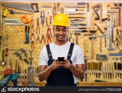 profession, construction and building - happy smiling indian worker or builder in helmet using smartphone over working tools on wall at workshop background. happy indian or builder in helmet using smartphone