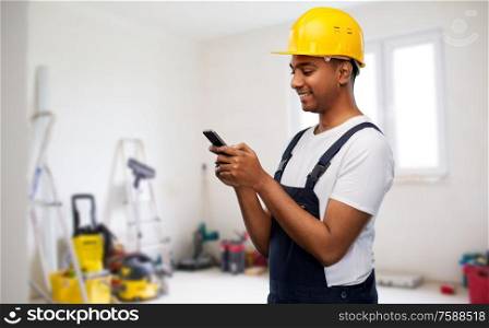 profession, construction and building - happy smiling indian worker or builder in helmet using smartphone over room with working equipment at new home or apartment background. happy indian or builder in helmet using smartphone