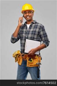 profession, construction and building - happy smiling indian worker or builder in helmet with clipboard calling on smartphone over grey background. indian builder in helmet calling on smartphone