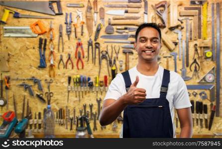profession, construction and building - happy smiling indian worker or builder showing thumbs up over working tools on wall at workshop background. happy indian worker or builder showing thumbs up