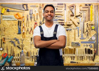 profession, construction and building - happy smiling indian worker or builder with crossed arms over working tools on wall at workshop background. happy indian worker or builder with crossed arms