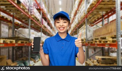 profession, job and people concept - happy smiling delivery woman in blue uniform with smartphone showing thumbs up gesture over warehouse background. delivery woman with smartphone showing thumbs up