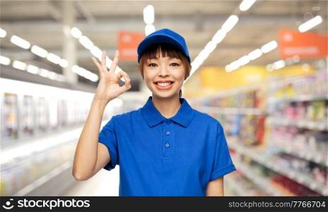 profession, job and people concept - happy smiling delivery woman or saleswoman in blue uniform showing ok gesture over supermarket background. happy smiling delivery woman showing ok gesture