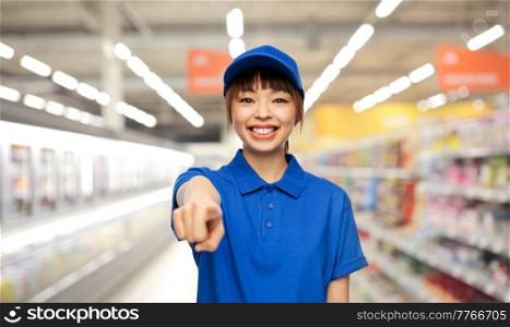 profession, job and people concept - happy smiling delivery woman or saleswoman in blue uniform pointing finger to camera over supermarket background. happy delivery woman pointing finger to camera