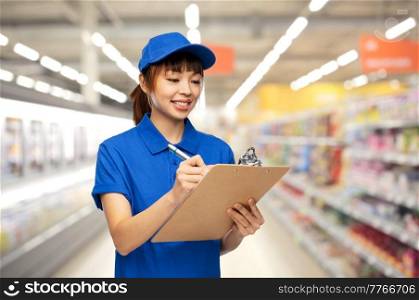 profession, job and people concept - happy smiling delivery woman or saleswoman in blue uniform with clipboard and pen over supermarket background. happy delivery woman with clipboard and pen