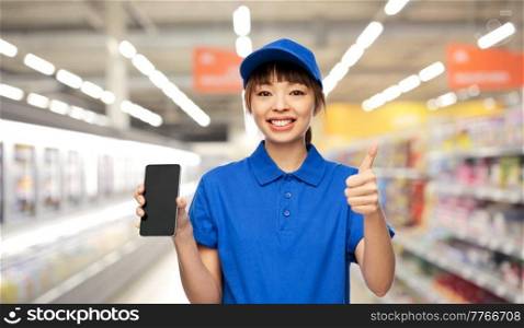 profession, job and people concept - happy smiling delivery woman or saleswoman in blue uniform with smartphone showing thumbs up gesture over supermarket background. delivery woman with smartphone showing thumbs up