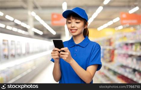profession, job and people concept - happy smiling delivery woman or saleswoman in blue uniform with smartphone over supermarket background. happy delivery woman with smartphone