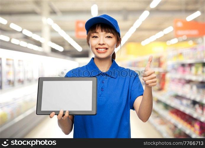 profession, job and people concept - happy smiling delivery woman or saleswoman in blue uniform with tablet pc computer showing thumbs up gesture over supermarket background. delivery woman with tablet pc showing thumbs up