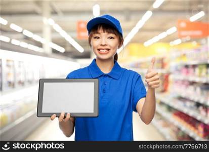 profession, job and people concept - happy smiling delivery woman or saleswoman in blue uniform with tablet pc computer showing thumbs up gesture over supermarket background. delivery woman with tablet pc showing thumbs up