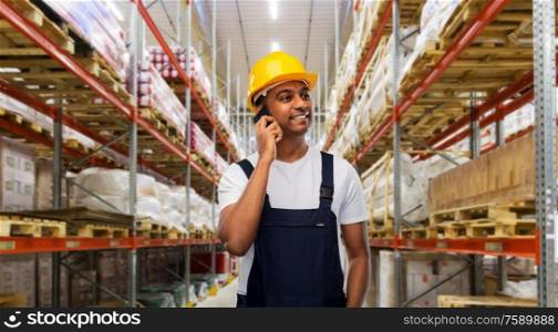 profession, logistic business and technology concept - happy smiling indian worker or loader in helmet calling on smartphone over warehouse background. indian worker calling on smartphone at warehouse