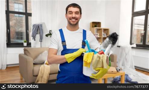 profession, service and people concept - happy smiling male worker or cleaner in overall and gloves with cleaning supplies over home room background. male cleaner with cleaning supplies at home
