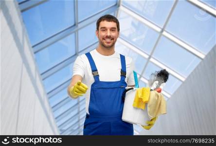 profession, service and people concept - happy smiling male worker or cleaner in overall and gloves with cleaning supplies pointing to camera over glasshouse background. male cleaner in overall with cleaning supplies