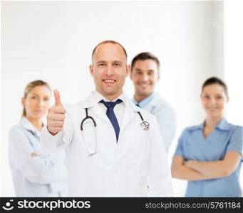 profession, teamwork, gesture and medicine concept - smiling male doctor with stethoscope in coat over group of medics showing thumbs up
