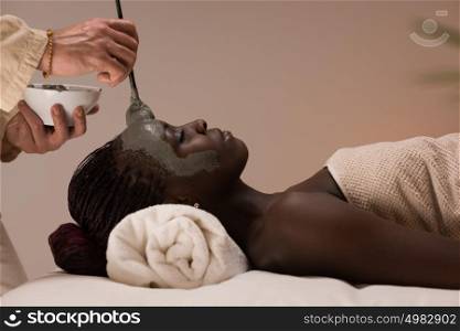 Professional beautician applying mud mask to african woman at spa salon