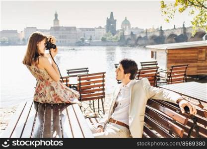 Professional beautiful female photographer makes photo of her husband who poses at camera on wooden bench at outdoor cafe near beautiful panoramic ancient city view. People and hobby concept