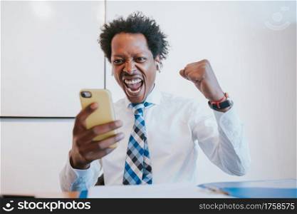 Professional businessman using reading good news on his mobile phone and celebrating victory while working at office. Business and success concept.