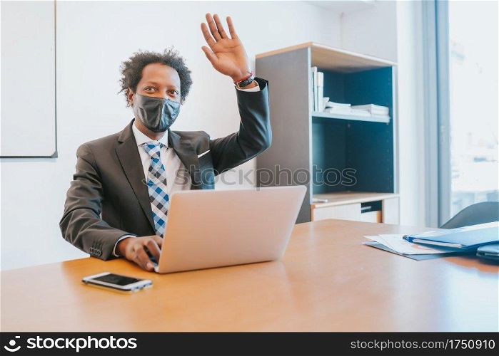 Professional businessman wearing face mask and saying hello to someone while working with his laptop at office. Business concept.