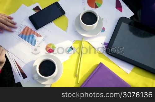 Professional businesspeople having a coffee break while working at the meeting outdoors, hands closeup top view with tablet, phone and paperwork on background