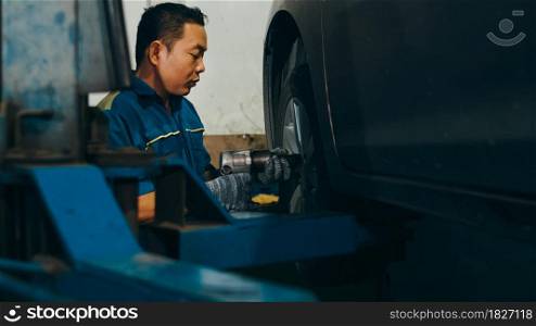 Professional car mechanic changing a car tire on lifted automobile at repair service station. Wheel alignment work at workshop night. Skillful Asian guy in uniform fixing car.