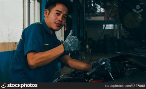 Professional car mechanic looking at camera and smiling at repair service station. Skillful Asian guy in uniform fixing car at mechanics garage at night. Car service maintenance.