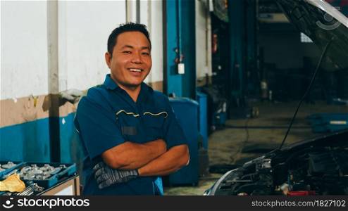 Professional car mechanic looking at camera and smiling at repair service station. Skillful Asian guy in uniform fixing car at mechanics garage at night. Car service maintenance.