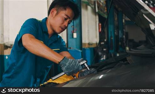 Professional car mechanic screwing details of car engine on lifted automobile at repair service station. Skillful Asian guy in uniform fixing car at mechanics garage at night.