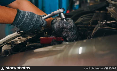 Professional car mechanic screwing details of car engine on lifted automobile at repair service station. Skillful Asian guy in uniform fixing car at mechanics garage at night.