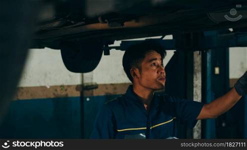 Professional car mechanic using paperwork makes the oil and engine check to the car on lifted automobile at repair service station night. Skillful Asian guy in uniform fixing car.
