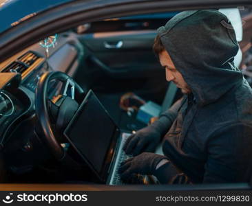 Professional car thief with laptop hacking security system, criminal lifestyle. Hooded male robber opening vehicle on parking. Auto robbery, automobile crime
