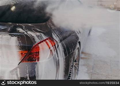 Professional car wash with high pressure washer and cleansing foam outdoors, close up of vehicle exterior surface in white soap suds getting wash at washing center. Professional car wash with high pressure washer and cleansing foam outdoors