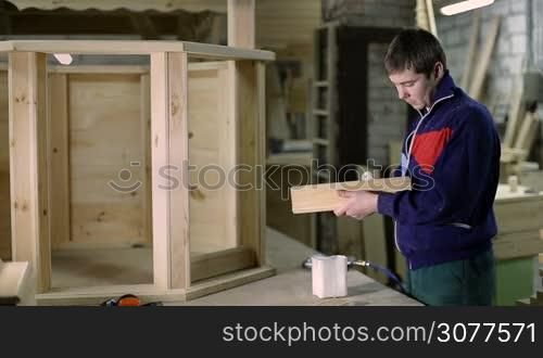 Professional carpenter gluing wooden surface with brush and constructing craft item using air nail gun in workshop. Carpenter working in woodwork shop.