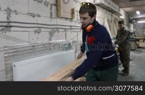 Professional carpenter measuring wooden plank with measuring tape in wood workshop. Joiner with protective goggles and earmuffs adjusting table saw to right size of board using measuring tape to cut.