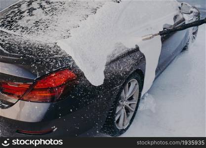 Professional carwashing and detailing. Covering black car with washing foam outdoors at self-service station, vehicle in white soap suds during professional auto cleaning with high-pressure gun. Car in white soap suds during professional auto cleaning outdoor
