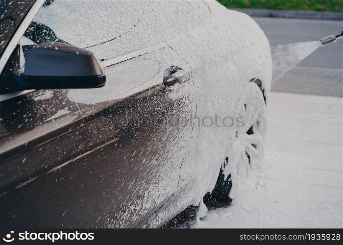 Professional carwashing with special detergents. Black luxury car in white snow foam during car wash outdoors, using high-pressure washer and soap to clean vehicle. Black luxury car in white snow foam during car wash outdoors