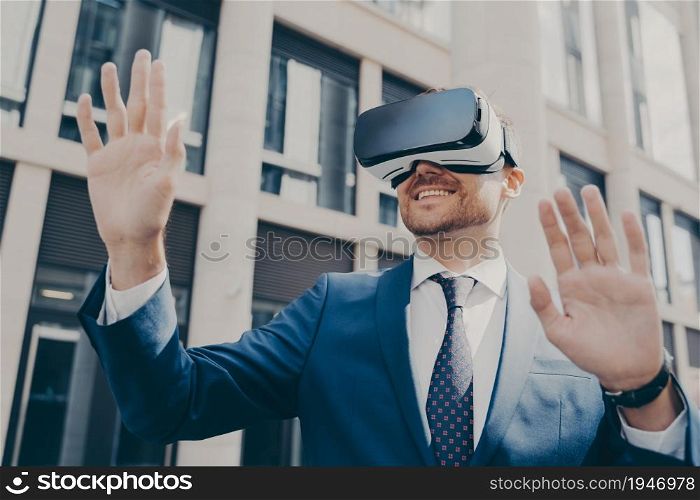 Professional excited office worker in blue suit standing outside alone and using VR glasses to visualize projects, holding something in virtual reality with his hands, building in background. Professional impressed worker standing outside alone and using VR glasses to visualize projects
