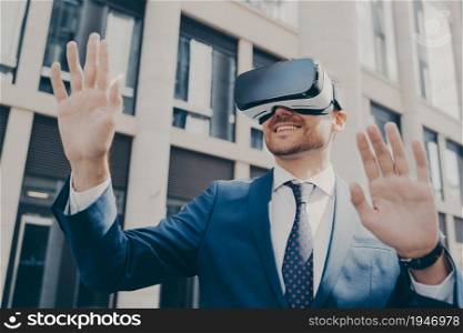 Professional excited office worker in blue suit standing outside alone and using VR glasses to visualize projects, holding something in virtual reality with his hands, building in background. Professional impressed worker standing outside alone and using VR glasses to visualize projects