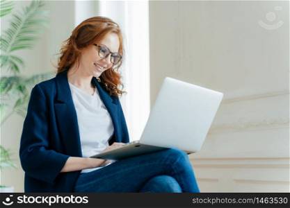 Professional female employer enjoys working process, sits crossed legs with laptop device, chats online, has positive look, has red curly hair, wears stylish elegant clothes. People and technology