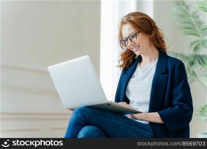 Professional female employer enjoys working process, sits crossed legs with laptop device, chats online, has positive look, has red curly hair, wears stylish elegant clothes. People and technology