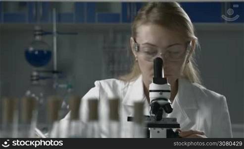 Professional female lab technician in protective eyewear doing microscope sample analysis and taking notes over modern lab enviroment. Life science female researcher microscoping in scientific genetic laboratory. Health care and biotechnology.