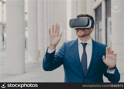 Professional male architect wearing augmented reality headset gesturing and smiling while standing outdoors, excited businessman using 3d goggles for work and business, moving virtual objects in air. Professional male architect in augmented reality headset gesturing while standing outdoors