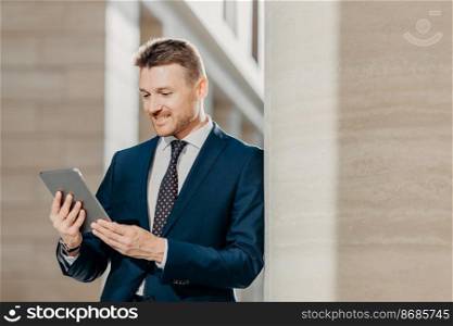 Professional male CEO in elegant suit, checks banking account on tablet in internet, stands in office hall, has pleased expression. Businessman updates application with financial news on website