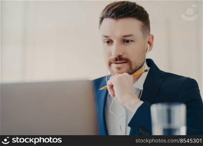 Professional male entrepreneur focused at laptop uses earphones listens necessary information watches webinar uses earphones holds pencil for writing down works online dressed in formal suit