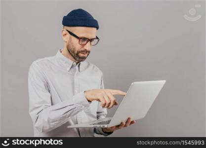 Professional male freelancer makes project work, points with index finger at screen of laptop computer, watches training webinar, holds modern gadget on hands, stands against grey studio wall