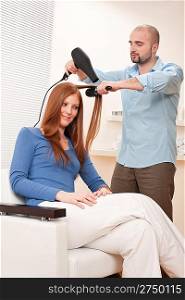 Professional male hairdresser with hair dryer and hair brush drying hair at salon with female red long hair customer