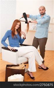 Professional male hairdresser with hair dryer and hair brush drying hair at salon with female red long hair customer