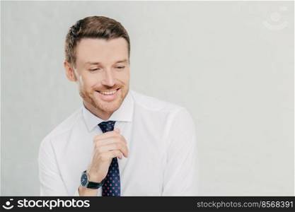 Professional male office manager giggles positively, concentrated down, shows white teeth, dressed in white shirt and tie, remembers something pleasant, isolated over white background with copy space