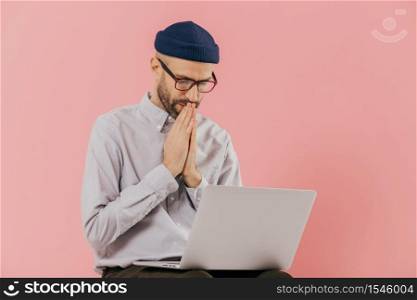 Professional male programmer keeps hans in praying gesture, looks at screen of laptop computer, believes in successful result of his work, wears formal shirt and hat, isolated over pink background
