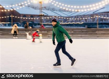 Professional male skater shows his skating talents, being sure on ice ring, looks delightfully in camera, demonstrates his nice skills. Smiling happy man on skating ring. Leisure and professionalism