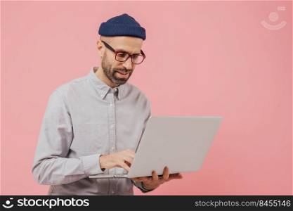 Professional male worker searches for interesting movie to watch, uses gadget, types information on laptop computer, wears formal clothes, isolated over pink background. Online communication